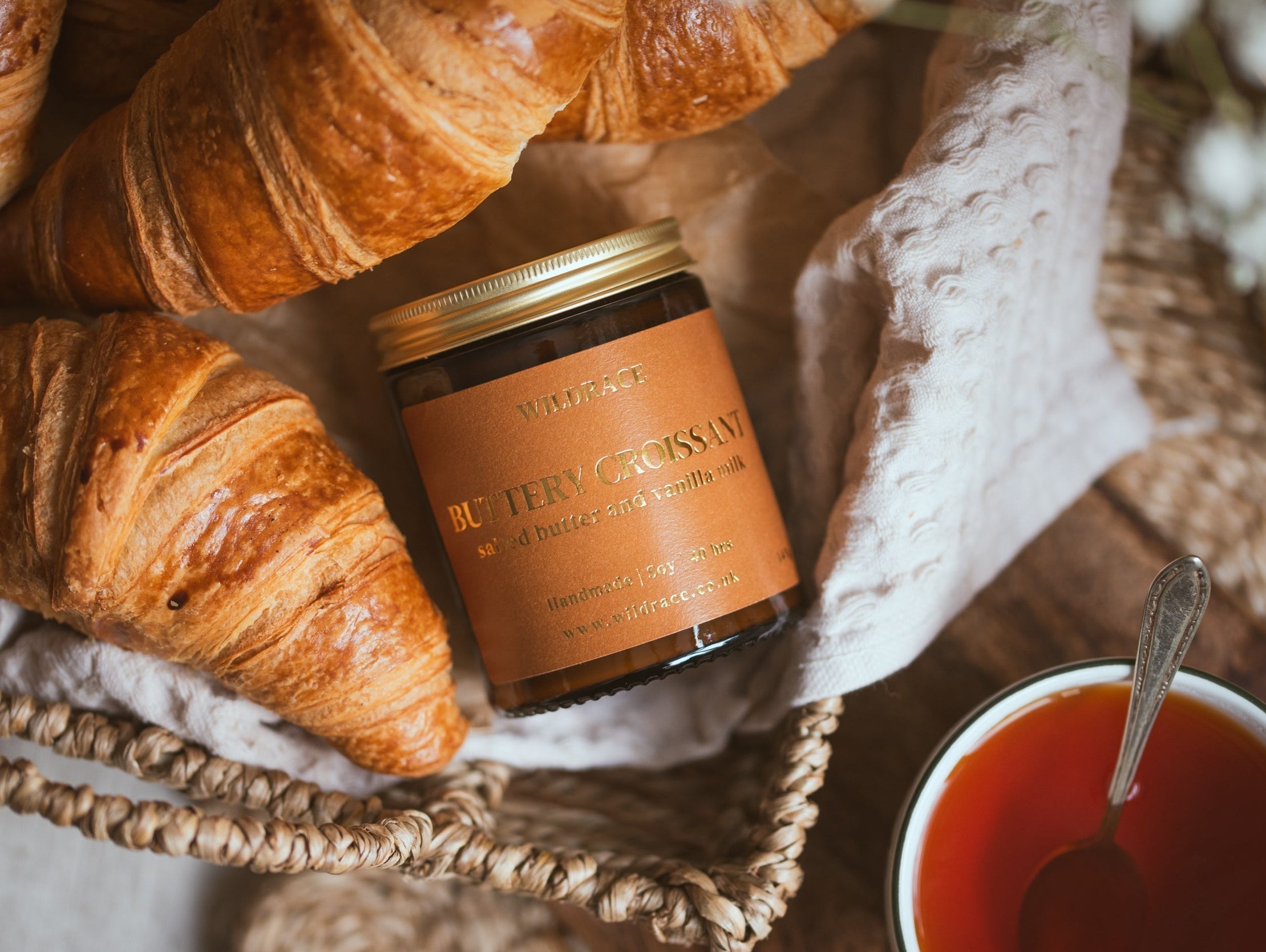 Wildrace Buttery Croissant candle in a woven basket surrounded by fresh croissants, with a cup of tea nearby.