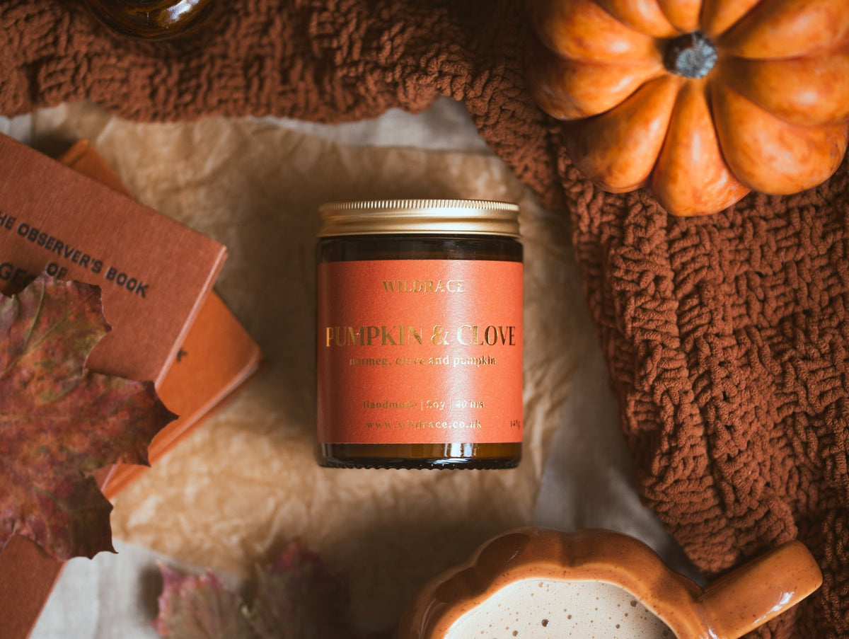 Wildrace Pumpkin & Clove candle with the lid on, styled flat lay with a vintage book, autumn leaf, pumpkin, and knit blanket.
