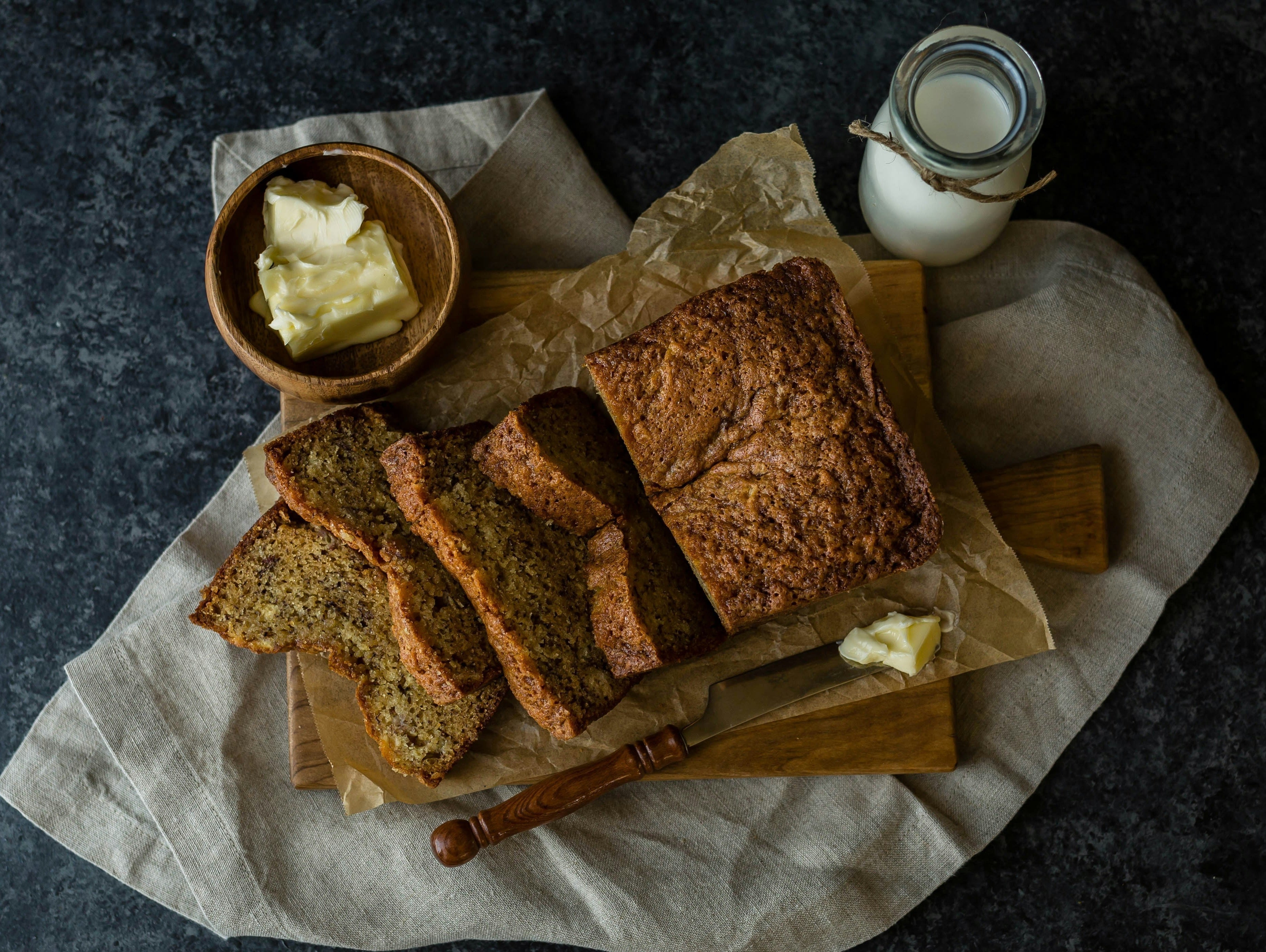 Sliced banana bread on a wooden board with a bowl of butter and a small jug of milk.