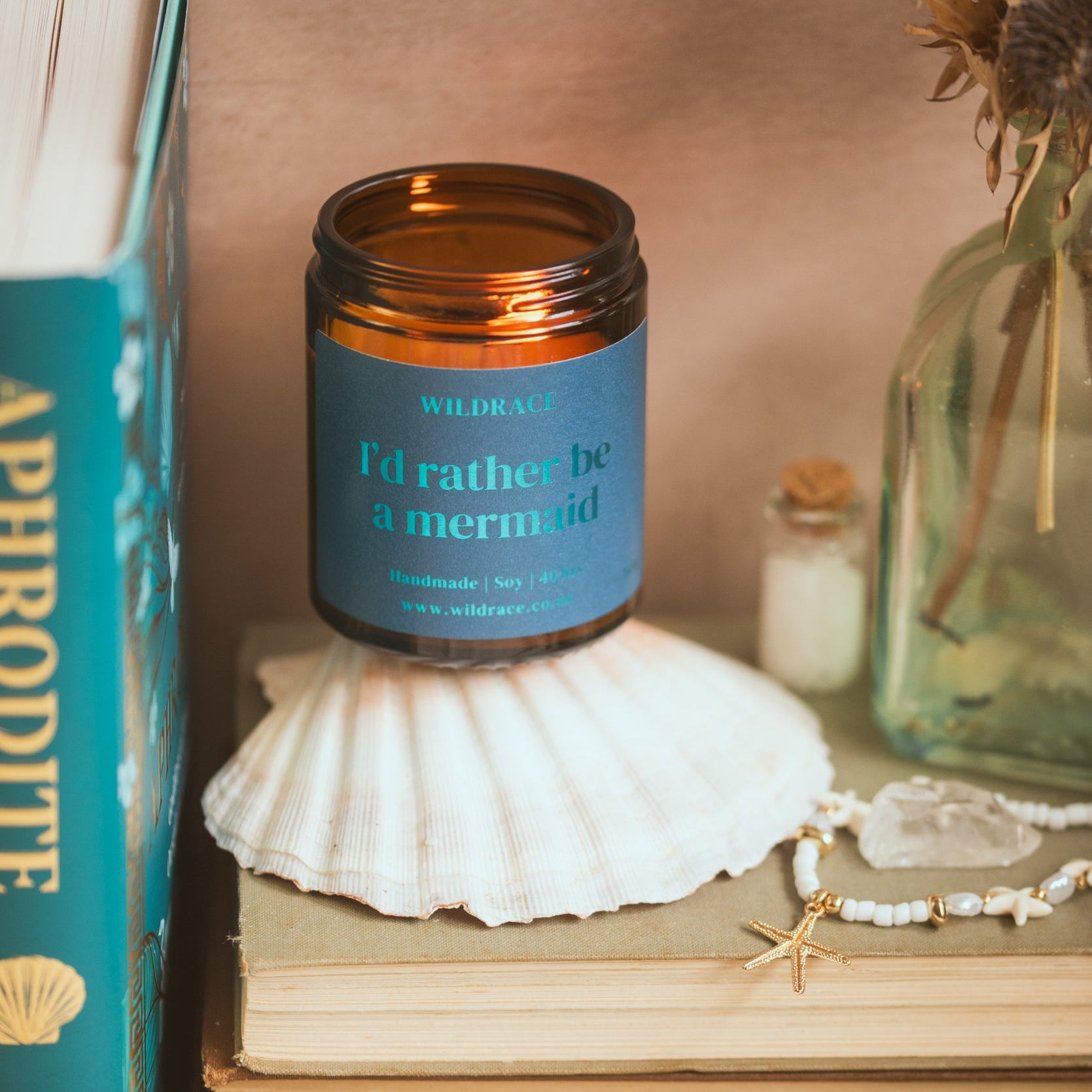Candle in a jar labeled 'I'd rather be a mermaid' on a stack of books with seashells and a plant.