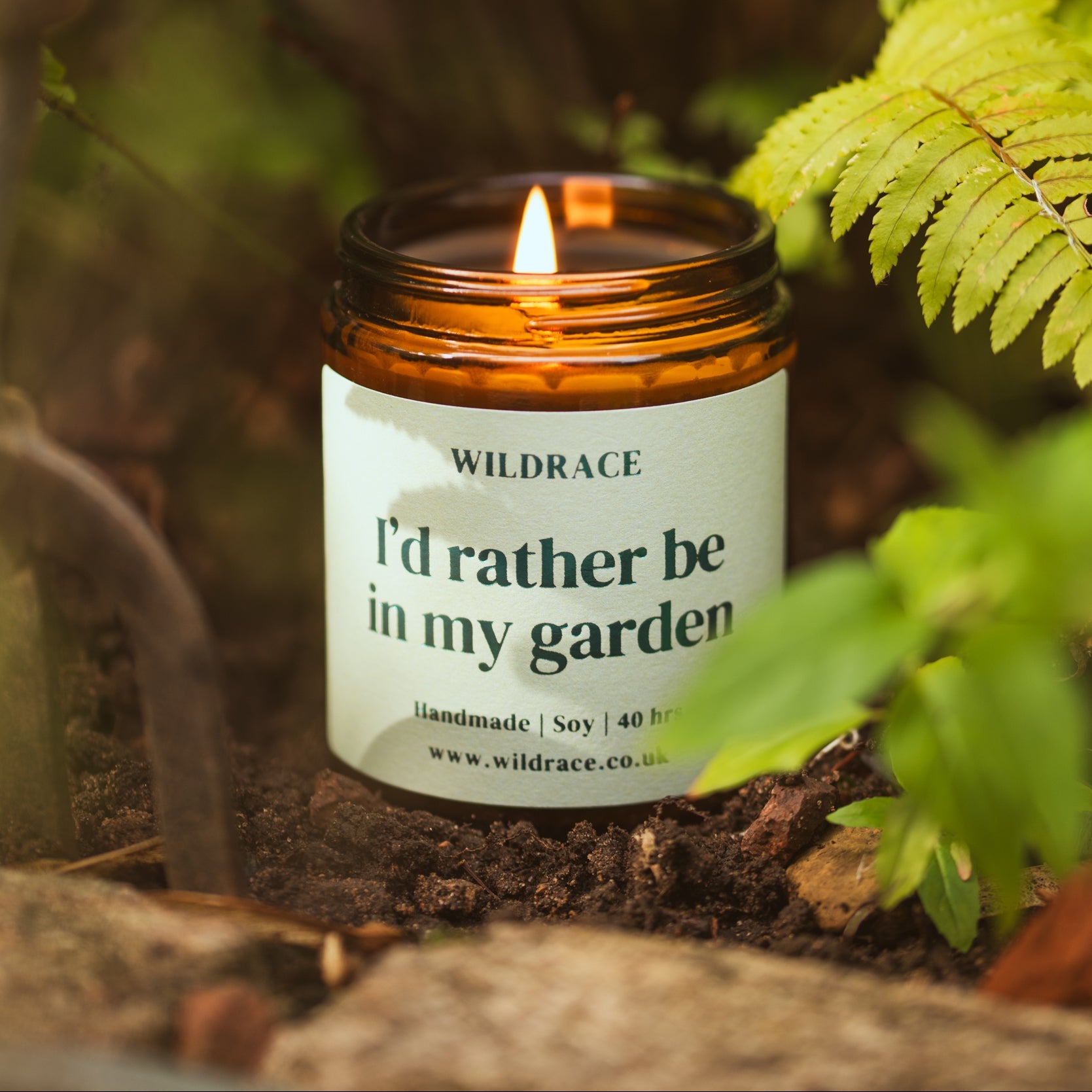 Candle in an amber jar with a label reading 'I'd rather be in my garden' surrounded by greenery.