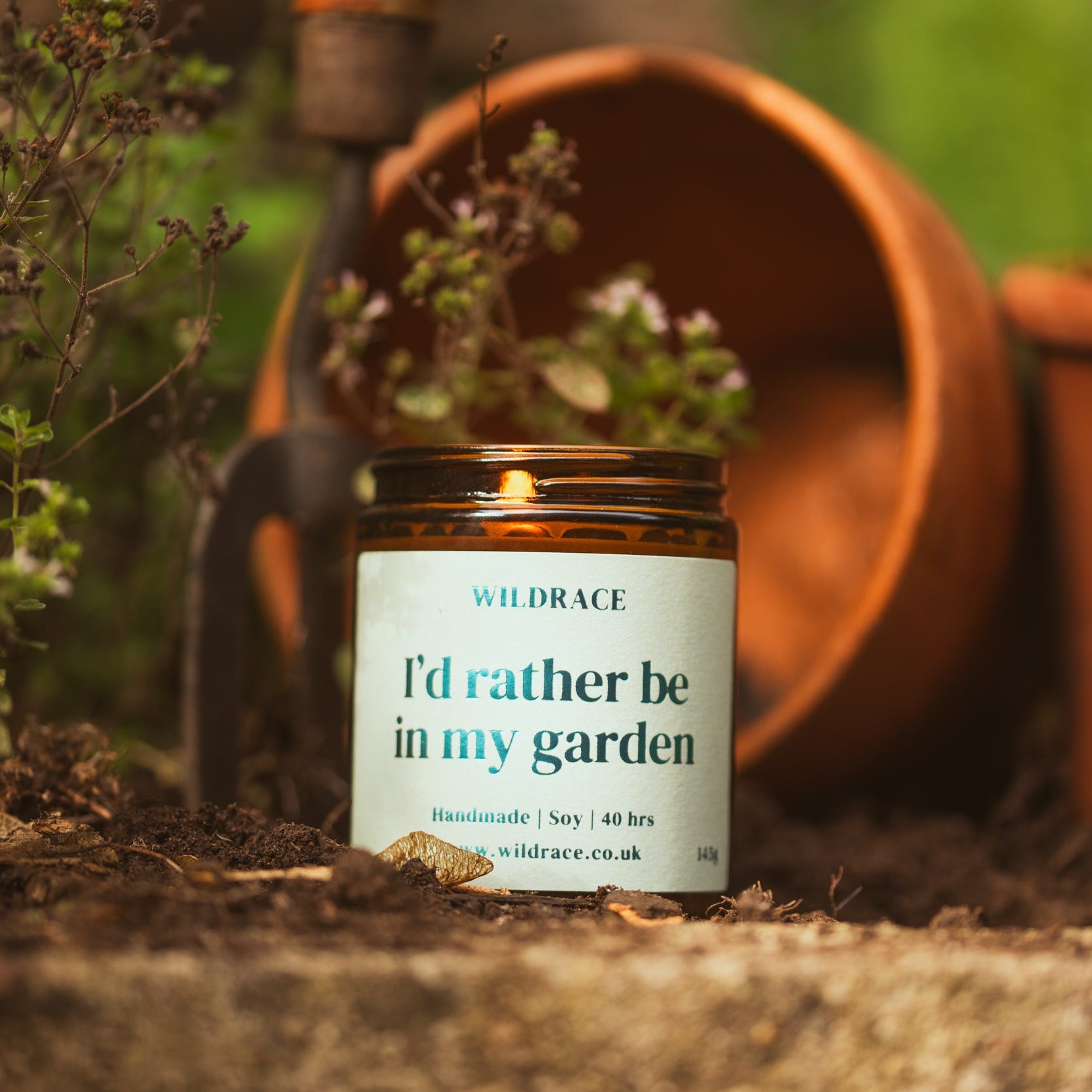 Candle labeled 'I'd rather be in my garden' with gardening tools and pots in the background