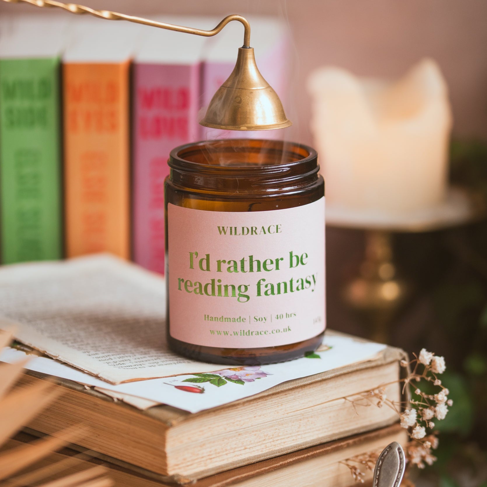 Candle labeled 'I'd rather be reading fantasy' on top of books with a cup of tea and spoon in the foreground.