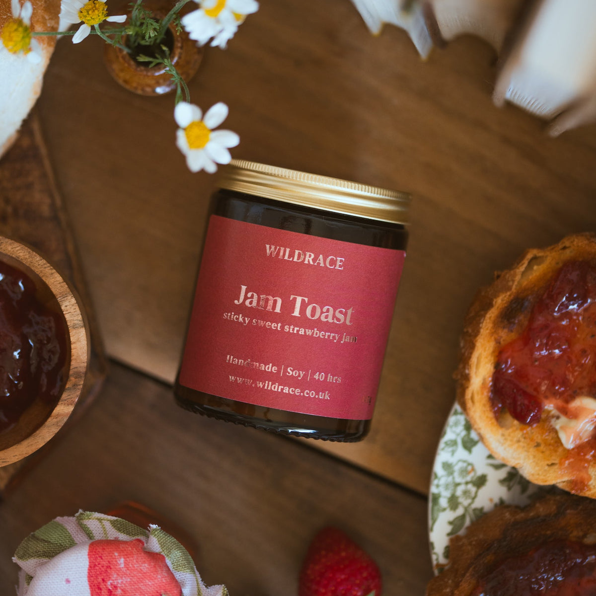 A Wildrace Jam Toast candle, on a wooden table surrounded by food props including strawberry jam on toast, strawberries, daisies, and a cup of tea.