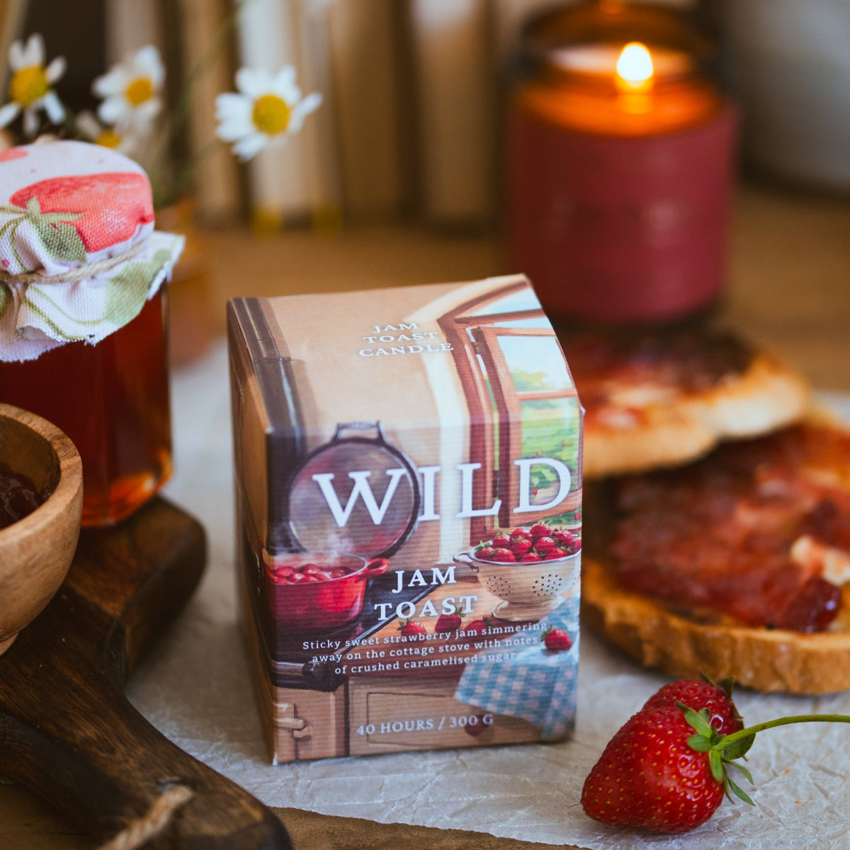 A box of Wildrace Jam Toast candle sits on a rustic wooden table surrounded by a lit candle, strawberries, toast, and a jar of jam in a warm, cosy setting.