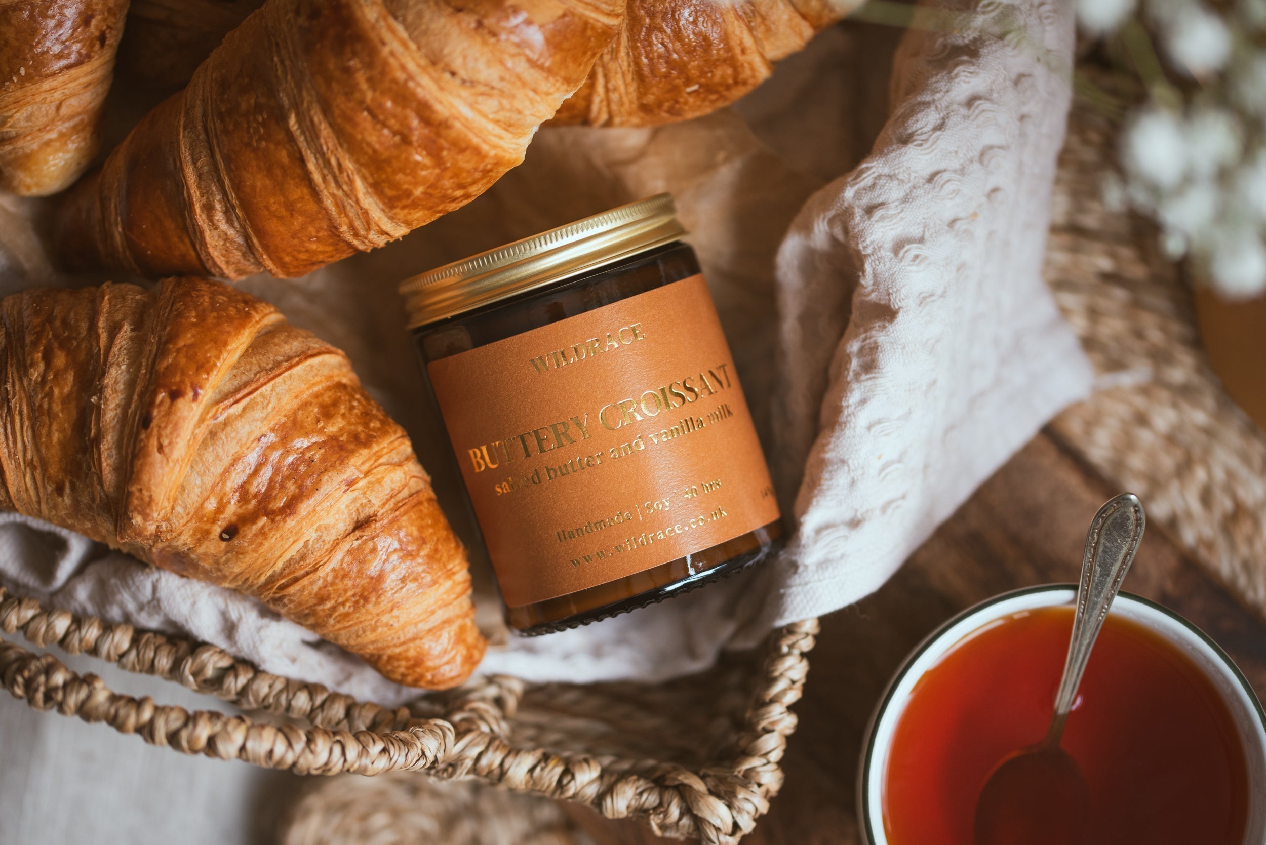 Wildrace Buttery Croissant candle in a woven basket surrounded by fresh croissants, with a cup of tea nearby.