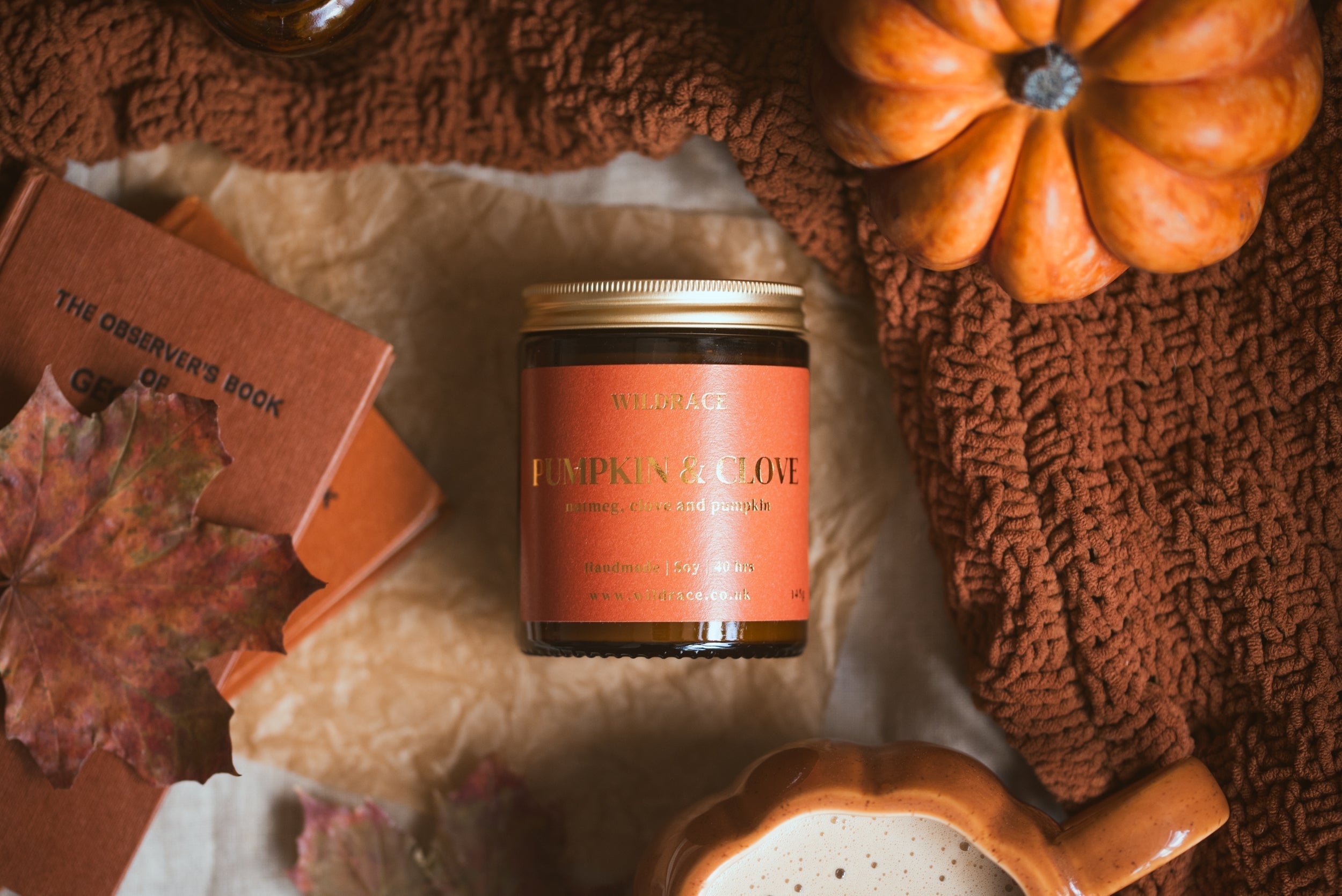 Wildrace Pumpkin & Clove candle with the lid on, styled flat lay with a vintage book, autumn leaf, pumpkin, and knit blanket.