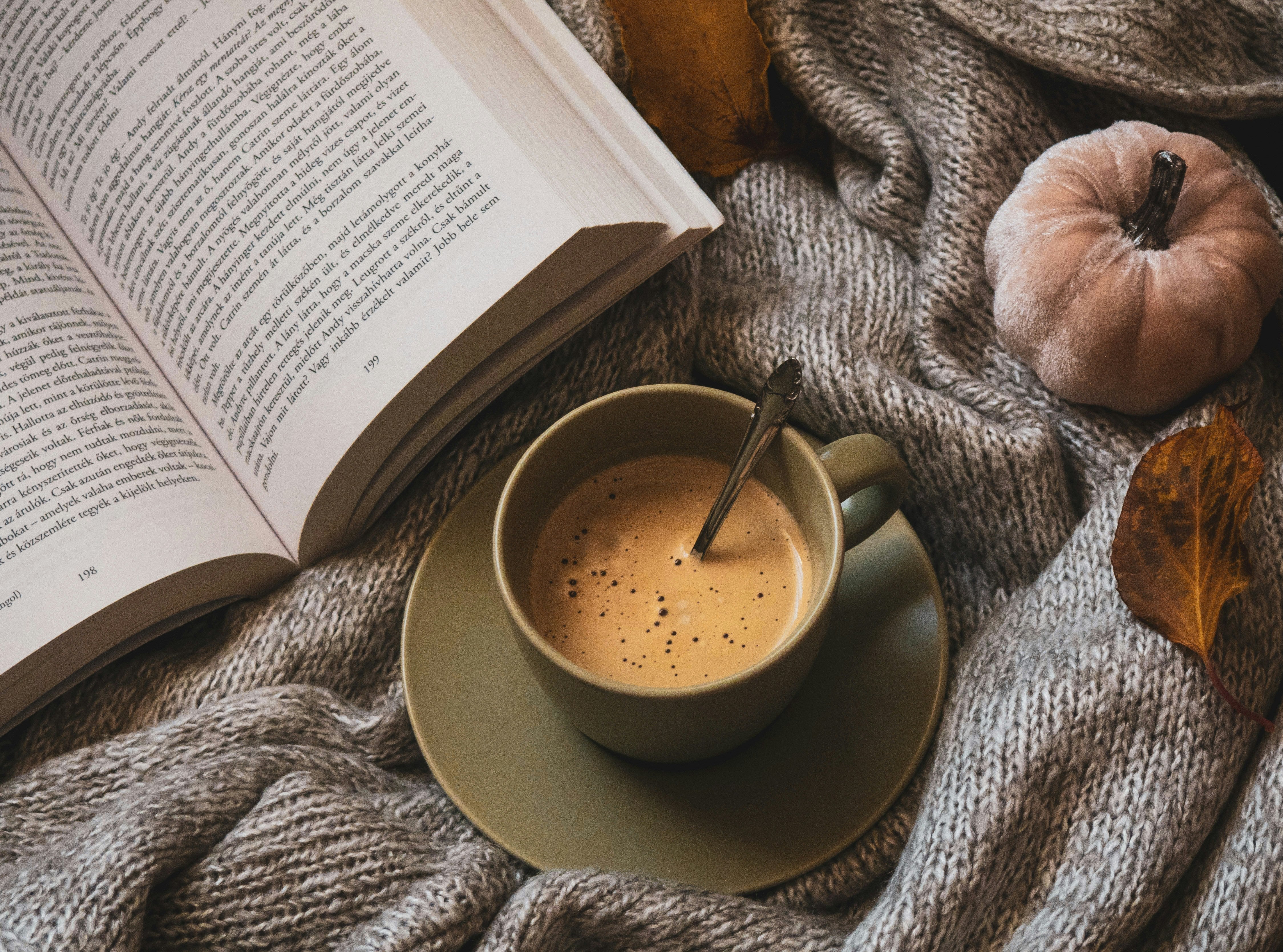 Cup of coffee on a knit blanket next to an open book, autumn leaf, and a small white pumpkin.