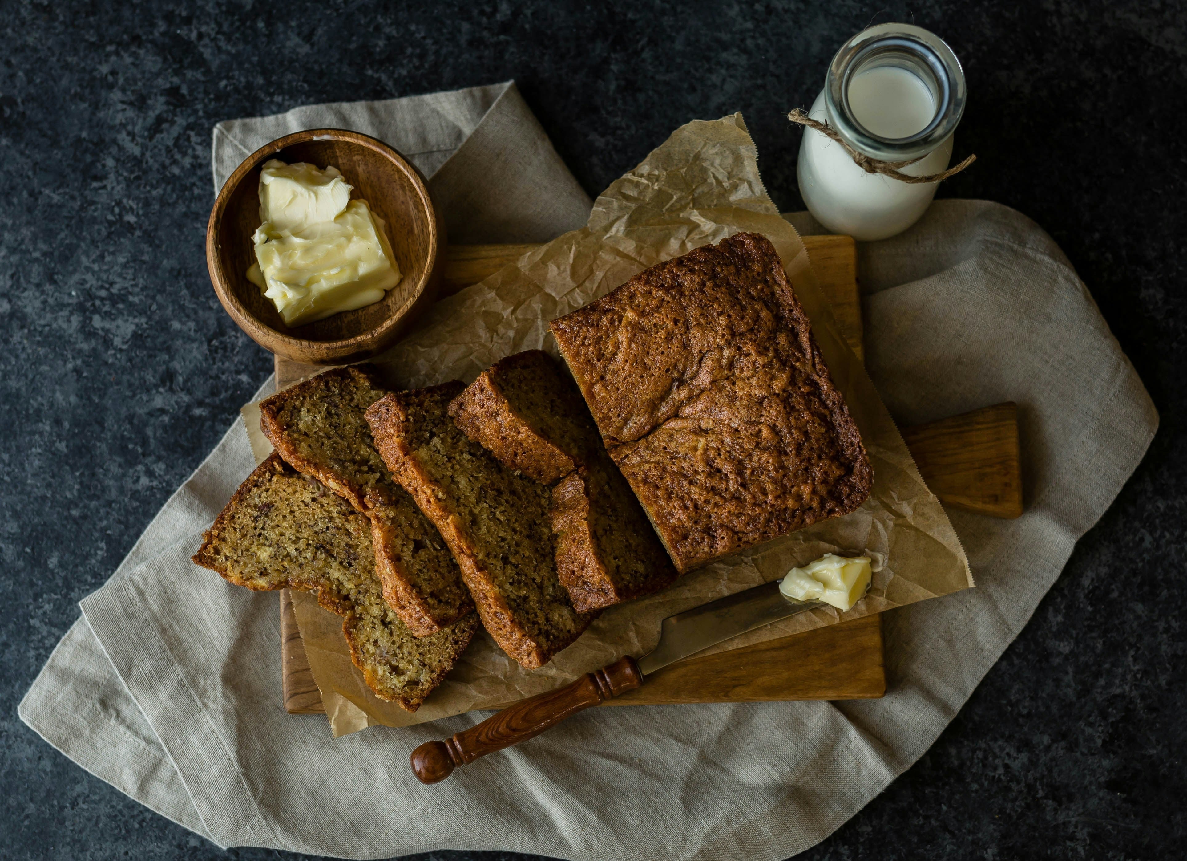 Sliced banana bread on a wooden board with a bowl of butter and a small jug of milk.