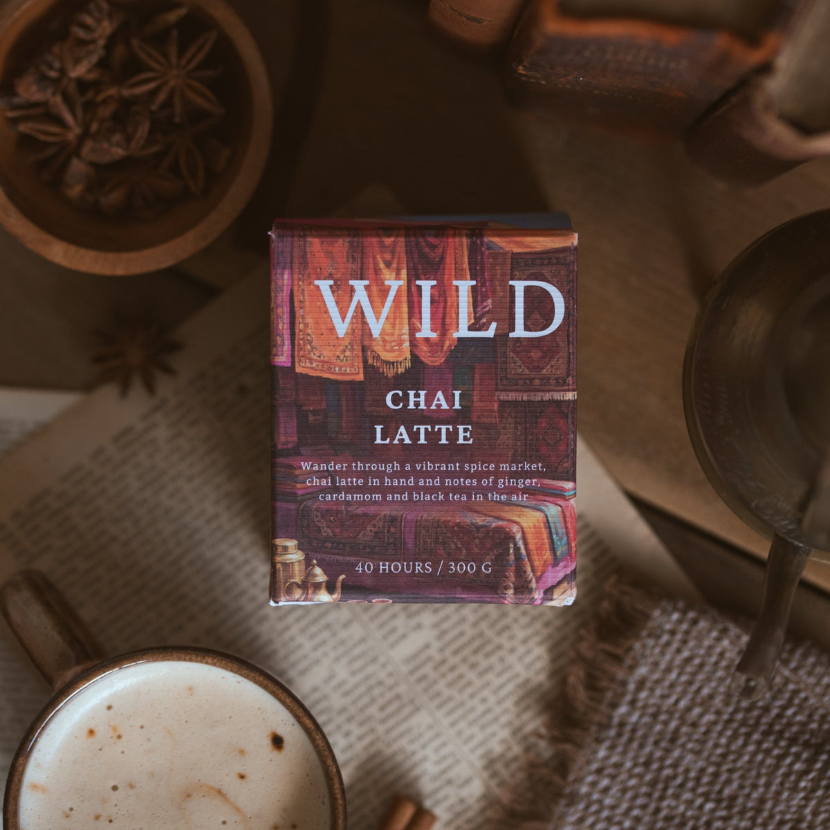 A WILDRACE Chai Latte candle is displayed in a cosy, rustic flat-lay setting with a cup of latte, cinnamon sticks, star anise, and old books in the background.