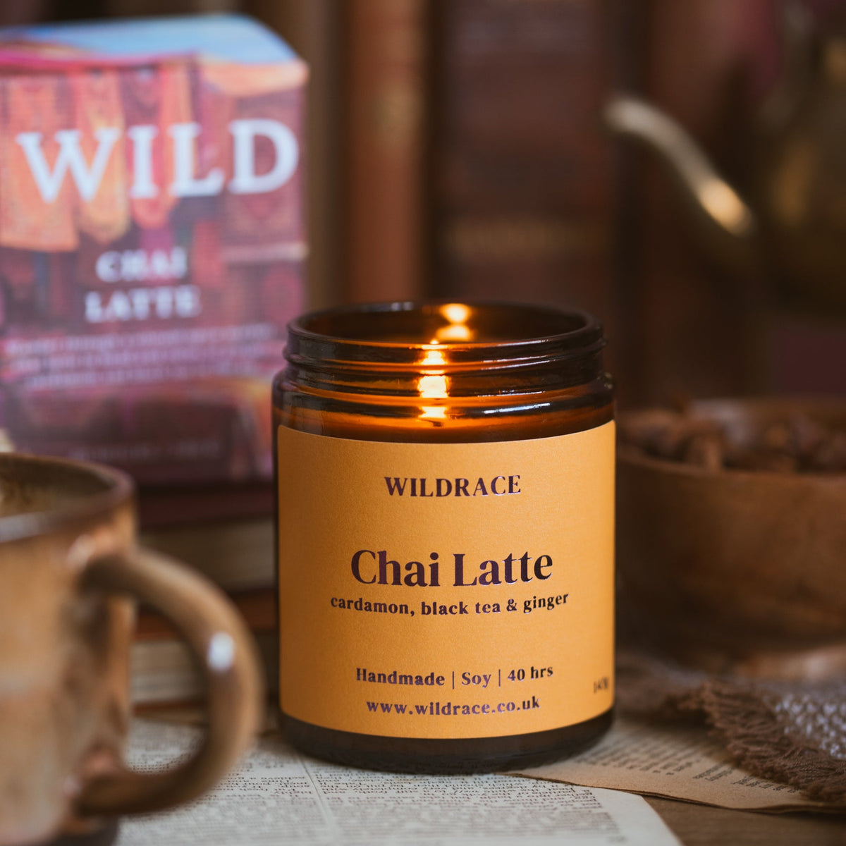 A lit amber glass jar candle labelled WILDRACE Chai Latte sits on a wooden surface with a warm orange label, surrounded by cosy props including a mug, book spines, and a bowl in a softly blurred indoor setting.