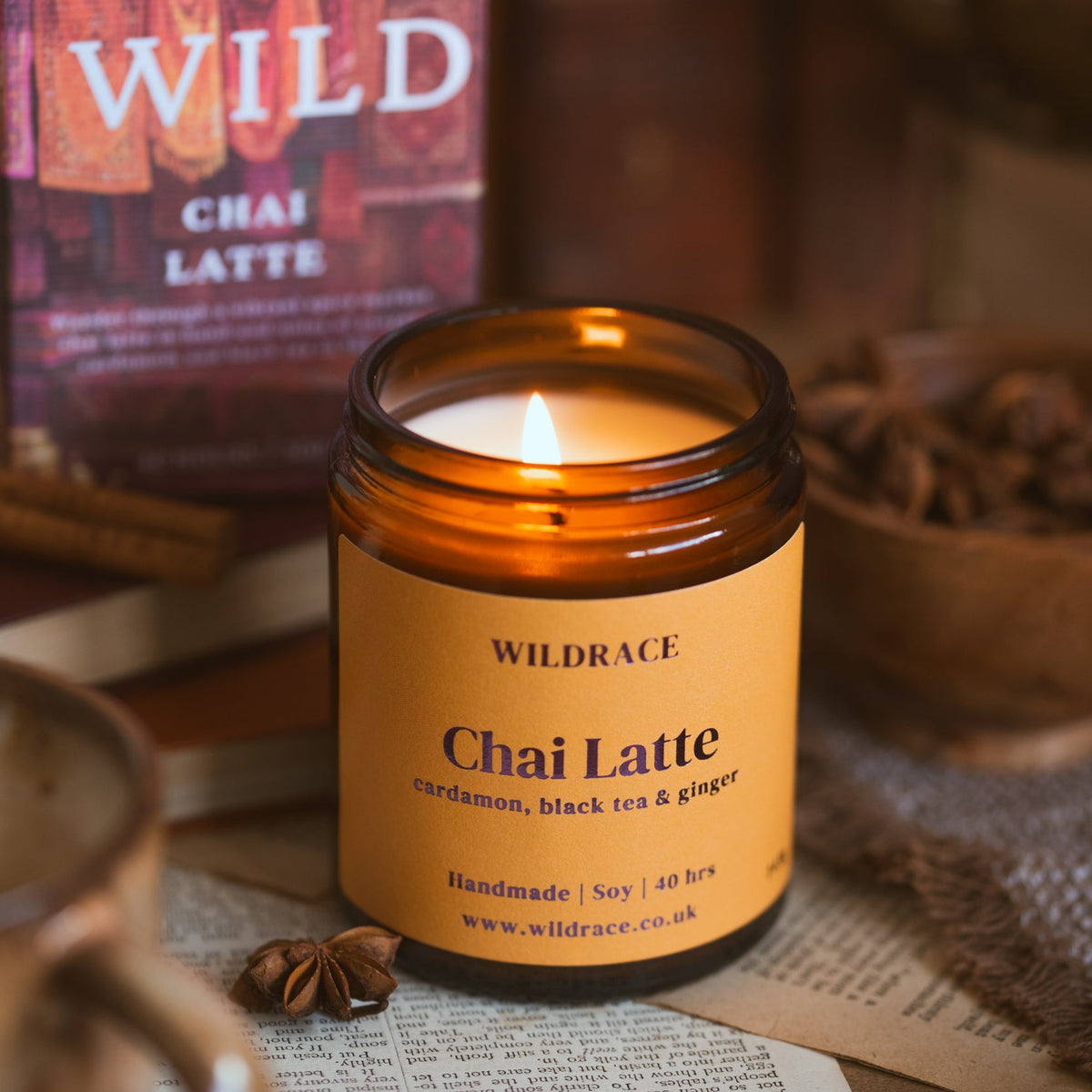 A lit amber glass jar candle labelled WILDRACE Chai Latte candle sits on a tabletop with a matching product box blurred in the background, surrounded by warm rustic props including books, a teapot, and star anise.