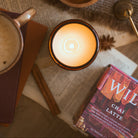 A cosy flat-lay scene featuring a lit candle in a dark amber glass jar, surrounded by star anise in a wooden bowl, a mug of coffee, a book, and a box of Wildrace Chai Latte tea on an open book.