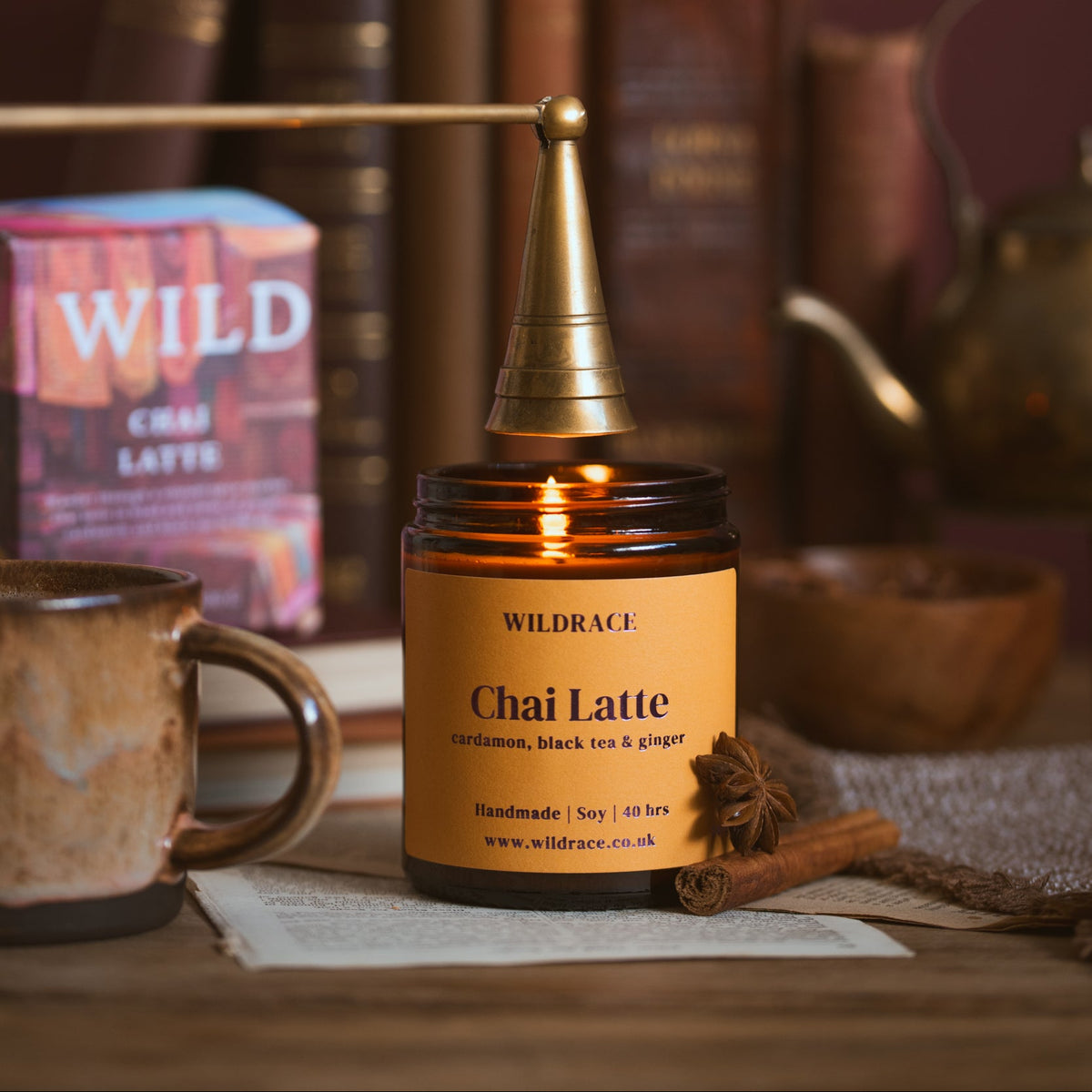A lit amber glass jar candle with a warm yellow label reading Wildrace Chai Latte, styled on a wooden surface with books, a mug, and a teapot in the background.