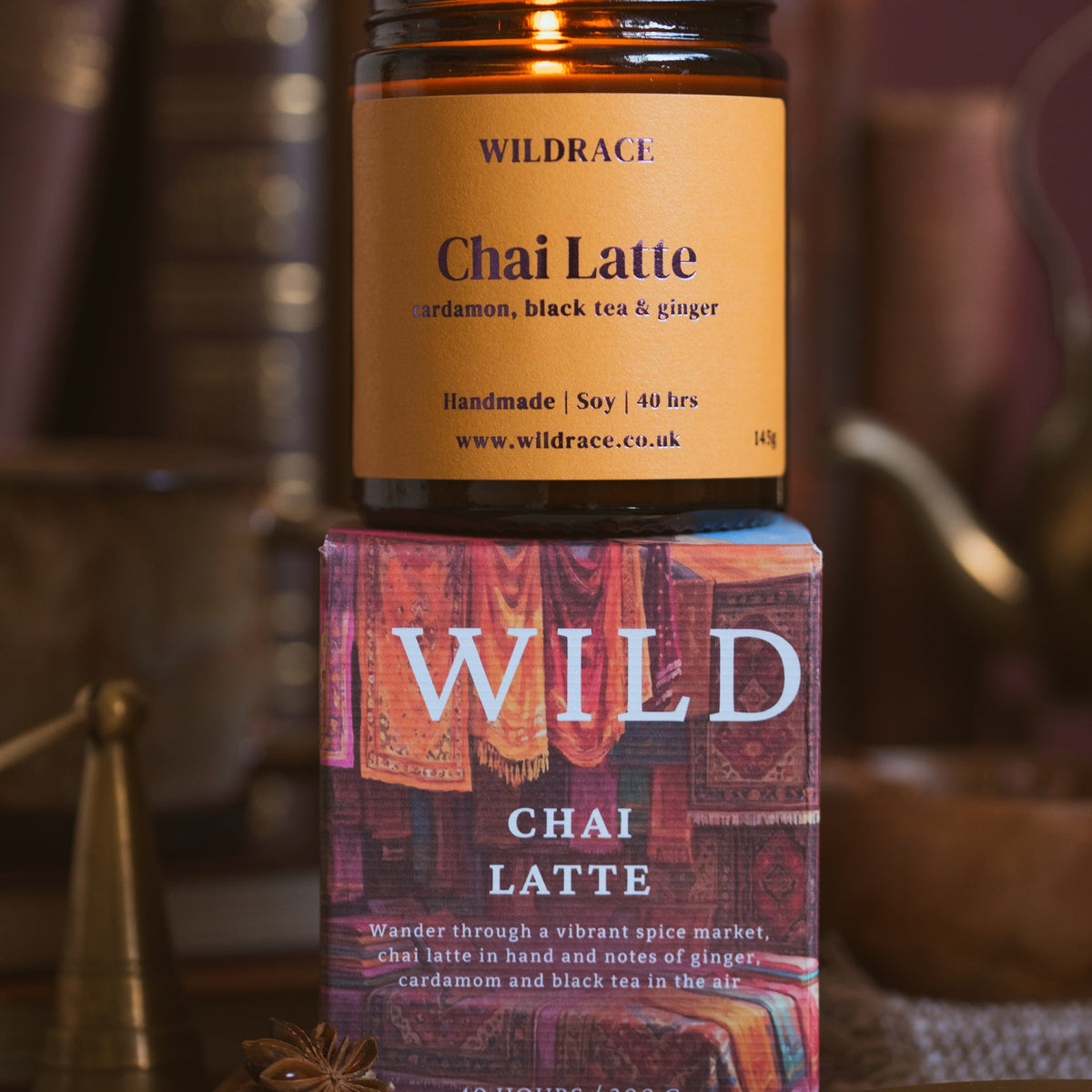 A Wildrace Chai Latte candle in a dark amber glass jar with a tan label, displayed on top of its colourful illustrated packaging box. The background is a warm, blurred rustic setting with books and a teapot.