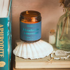 Candle in a jar labeled 'I'd rather be a mermaid' on a stack of books with seashells and a plant.