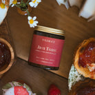 A Wildrace Jam Toast candle, on a wooden table surrounded by food props including strawberry jam on toast, strawberries, daisies, and a cup of tea.