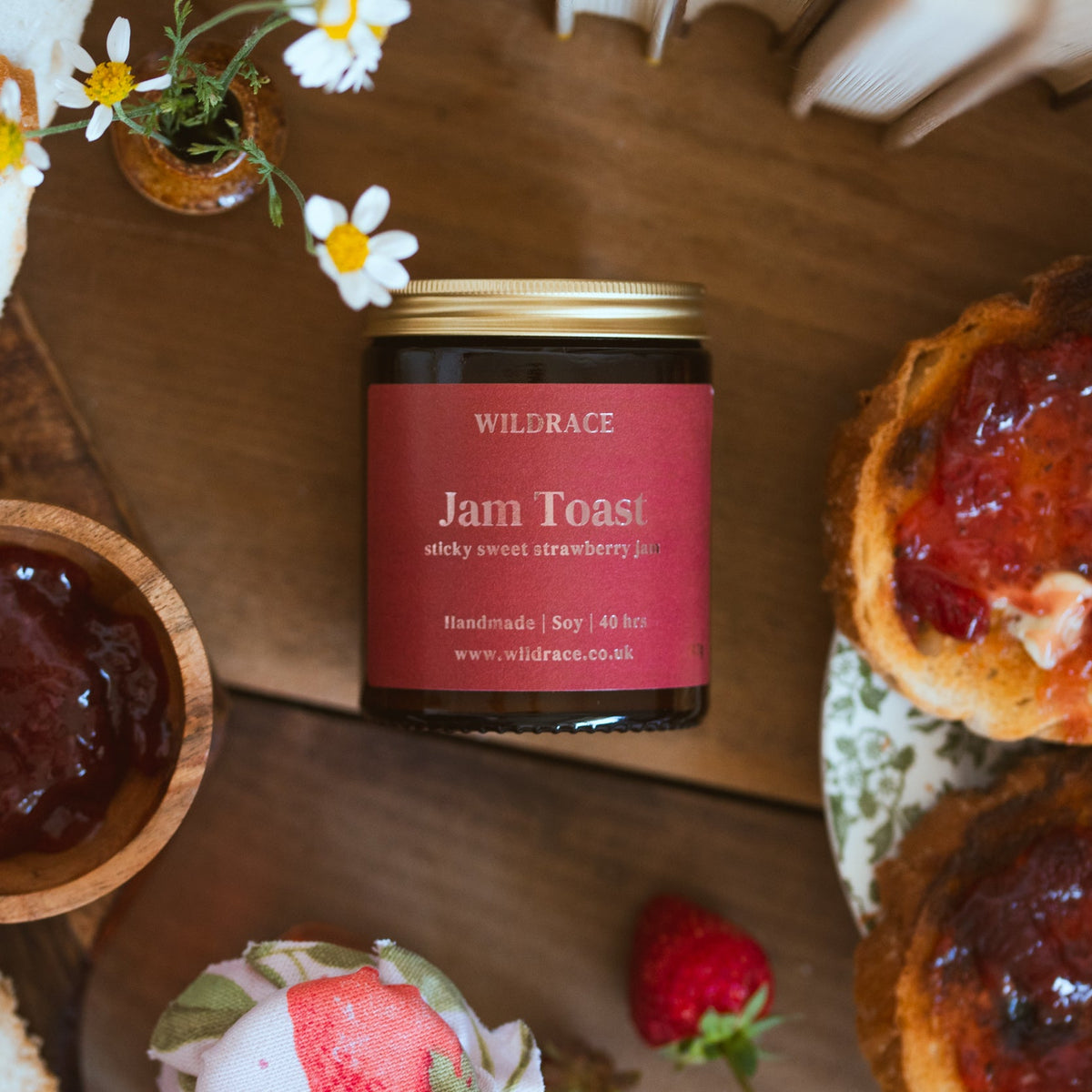 A jar of Wildrace Jam Toast candle, surrounded by bread, a small bowl of jam, strawberries, and daisies on a wooden table.