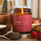 A lit amber glass jar candle with a redlabel reading 'WILDRACE Jam Toast' and 'sticky sweet strawberry jam,' styled on a tabletop with strawberries, bread, and a blurred background of books.