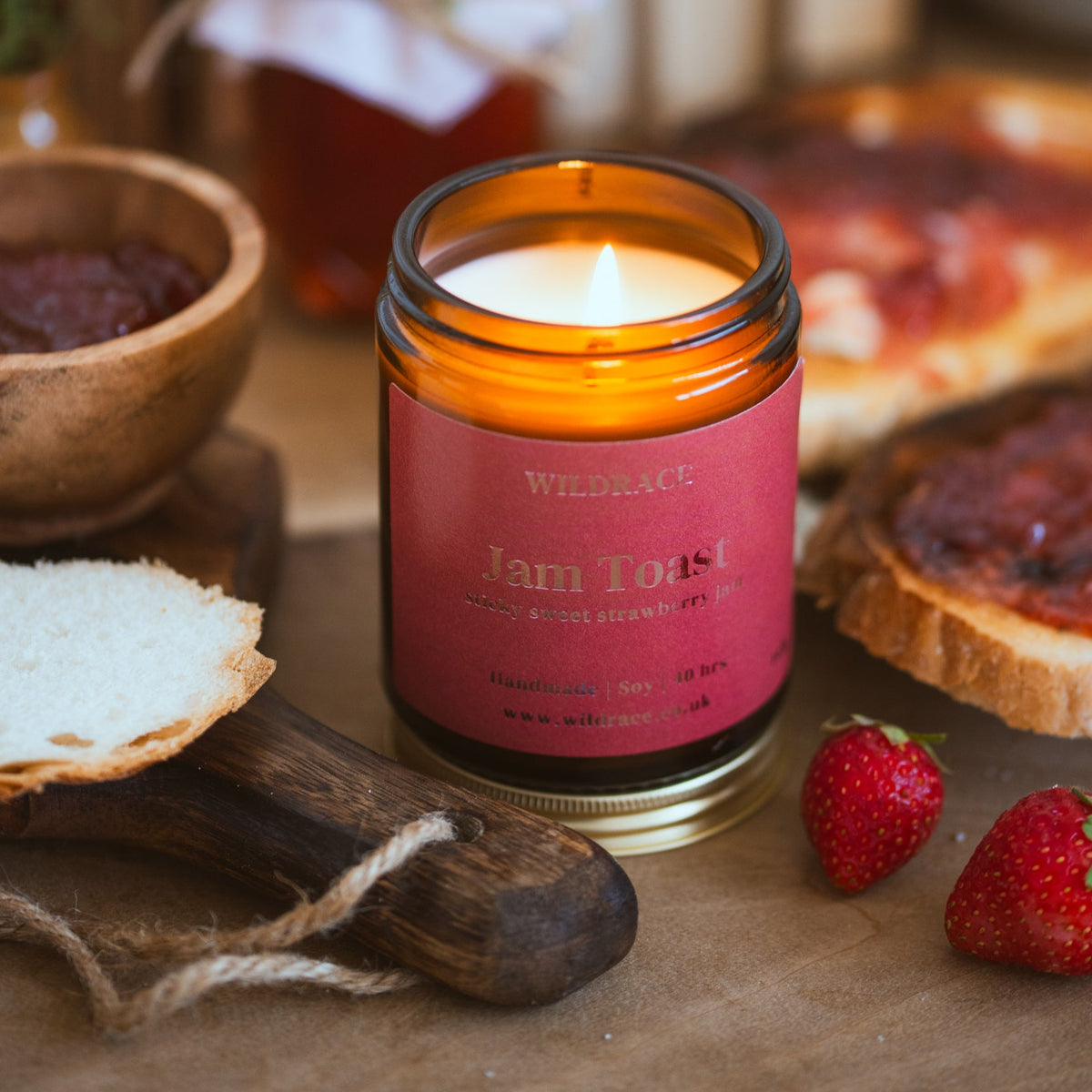 A lit amber glass jar candle with a red label reading 'Wildrace Jam Toast' sits on a wooden surface surrounded by toast, strawberries, and a bowl of jam in a warm, rustic setting.