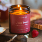 A lit Wildrace 'Jam Toast' soy candle in a dark amber glass jar, styled on a rustic wooden surface with a fresh strawberry and slices of toast in a warm, cosy setting.