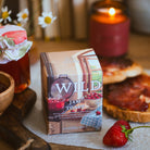 A box of Wildrace Jam Toast candle sits on a rustic wooden table surrounded by a lit candle, strawberries, toast, and a jar of jam in a warm, cosy setting.
