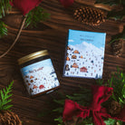 Candle and packaging with holiday design on a wooden surface with greenery and pine cones.